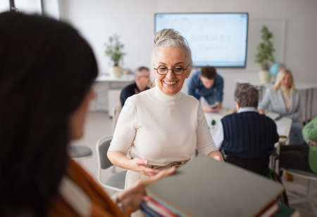 Happy senior woman student giving books to teacher in classroom.の写真素材