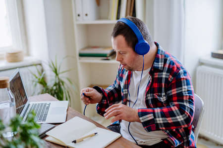 Young man with Down syndrome sitting at desk in office and using laptop, listening to music from headphones.の写真素材