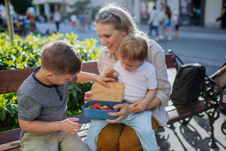 Young mother with little kids sitting on bench in city in summer, eating fruit snack and drinking water.の写真素材