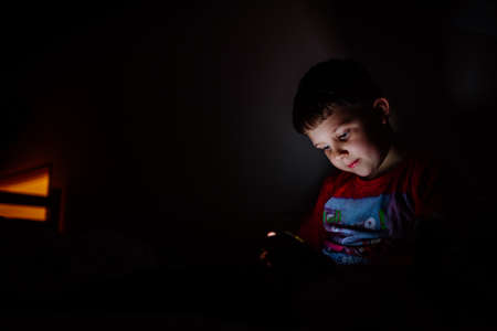Addicted little boy playing games on phone in bed at night.