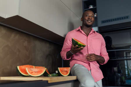 Multiracial man eating watermelon in his kitchen during hot sunny days.の写真素材