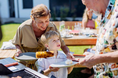 Grandparents giving grilled cheese and corn their grandson at garden grill party.の写真素材