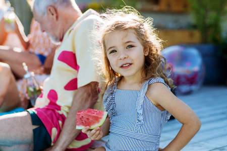 Cute little girl in swimsuit eating watermelon corn and sitting next to pool with family at garden party.の写真素材
