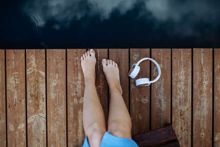 Close-up of womans legs and headphones lying on a pier by natureal lake in summer Top view.の写真素材