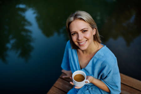 Happy young woman sitting by pond near cottege and enjoying cup of morning coffee on summer vacation in mountains. High angle view.の写真素材