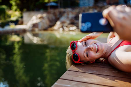 Rlaxed woman relaxing on the pier, lying and taking selfie near the natural lake in summer
