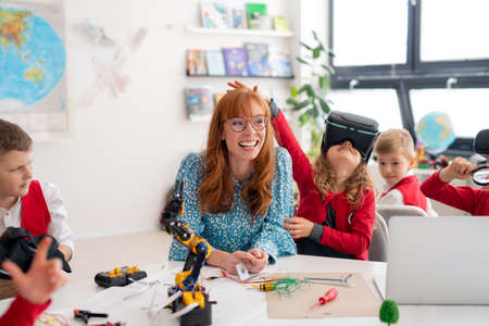 Happy schoolgirl wearing virtual reality goggles at class, having fun with teacher and classmates.