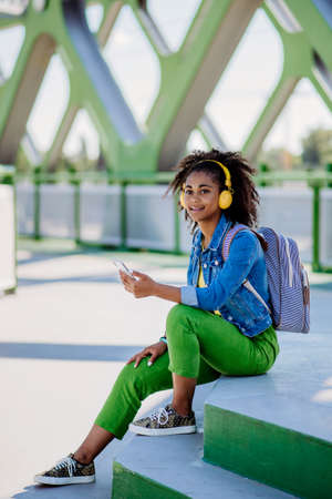 Multiracial teenage girl with backpack, headphones and smartphone resting after school in city...