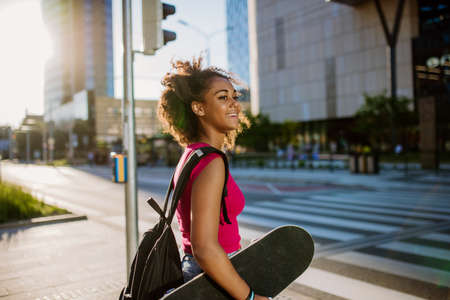 Multiracial teenage girl with backpack and skateboard, walking in city during summer day.の写真素材