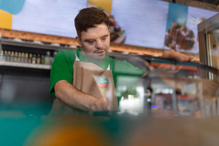 Happy waiter with Down syndrome standing by counter and prepairing hot-dog to a customer in cafe at...