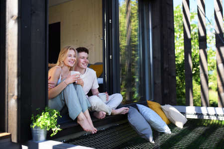 Young couple sitting with cup of coffee and cuddling in terrace in their new home in tiny house in woods, sustainable living concept.の写真素材
