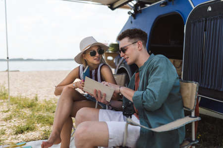 Young couple sitting together in front of van, camping and reading book.