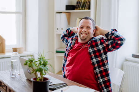 Young man with Down syndrome sitting at desk in office and using laptop, looking at camera and smiling.の写真素材