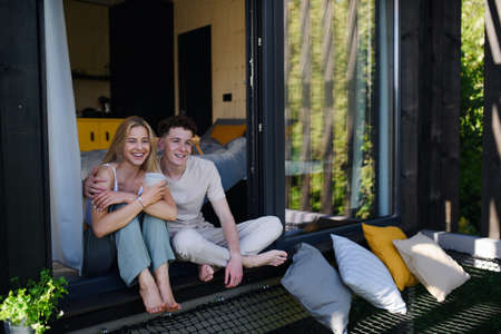 Young couple sitting with cup of coffee and cuddling in terrace in their new home in tiny house in...