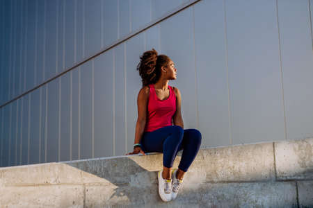 Young multiracial girl in sportswear resting and sitting after jogging in city.