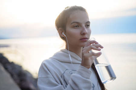 Young beautiful sportive girl listening to music and drinking water at sunrise by the lake.