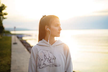 Young beautiful sportive girl listening to music at sunrise by the lake.の写真素材