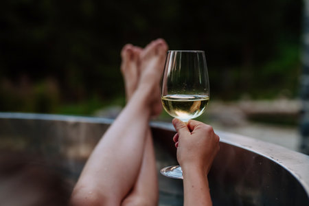 Unrecognizable young woman with feet up relaxing with glass of wine in hot tub outdoor in nature.の写真素材