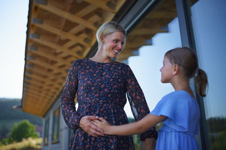 Little daughter stroking pregnant belly his mother, in front of their new wooden house.の写真素材