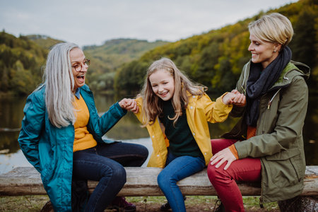 Small girl with mother and grandmother sitting on bench and having fun outoors by lake.の写真素材