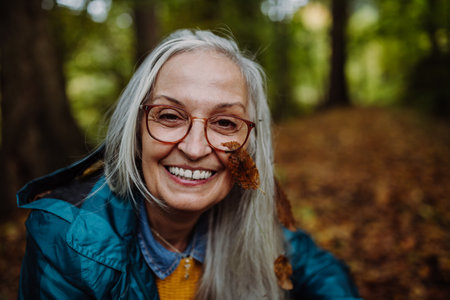 Portrait of happy senior woman with leaf on her cheek on walk outdoors in forest in autumn.