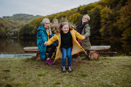 Small girl having fun, her mother and grandmother sitting on bench outoors by lake.の写真素材