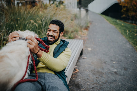 Happy young man training and cuddling with his dog outdoors in city park, during cold autumn day.の写真素材