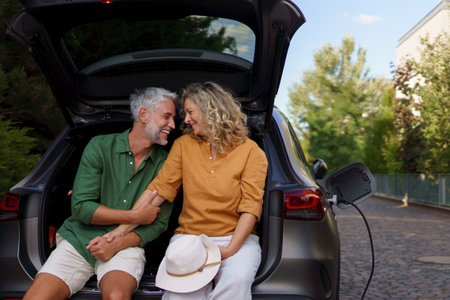 Middle-aged couple sitting in trunk while waiting for charging car before travelling on summer holiday.の写真素材