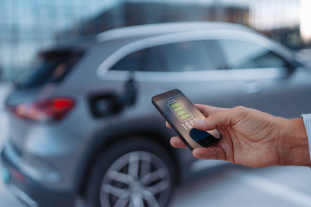 Man holding smartphone while charging car at electric vehicle charging station.