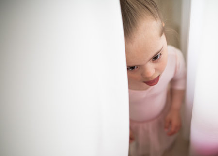 High angle view of little girl with down syndrome hidding behind curtain, at ballet class in dance studio. Concept of integration and education of disabled children.の写真素材