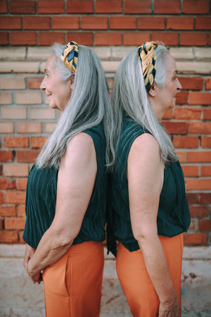 Side view of senior women, twins,in same clothes standing back to back, outdoor in city.の写真素材