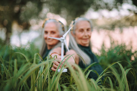 Senior women, twins holding plastic model of wind turbine in nature, concept of future, ecology and...