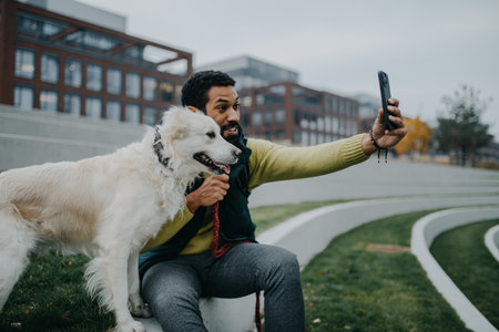 Happy young man resting and taking selfie with his dog outdoors in city park, during cold autumn day.の写真素材