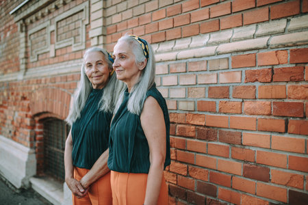 Senior women twins,in same clothes standing and posing in front of brick wall, outdoor in city.