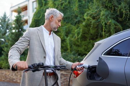 Close up of businessman in suit on way to work standing at eletric scooter and charging his electric car. Concept of eco commuting and green transportation.の写真素材