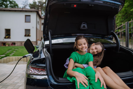 Happy sisters sitting in car trunk and waiting for electric car charging.の写真素材