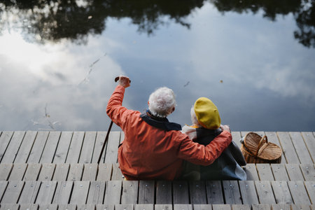 Happy senior couple at autumn walk near the lake, having break, sitting at pier.の写真素材