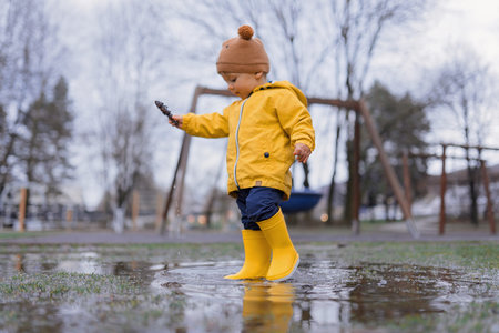 Happy little boy in yellow raincoat jumping in puddle and playing with conifer cone after rain in autumn day.の写真素材