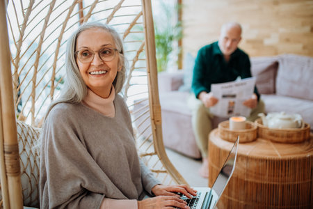 Senior woman sitting in outdoor swing and working at laptop, her husband resting at sofa. Concept of...