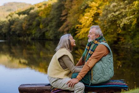 Senior couple in love sitting together on bench near lake, during autumn day.の写真素材