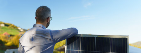 Rear view of businessman holding solar panel, standing outdoor at garden.