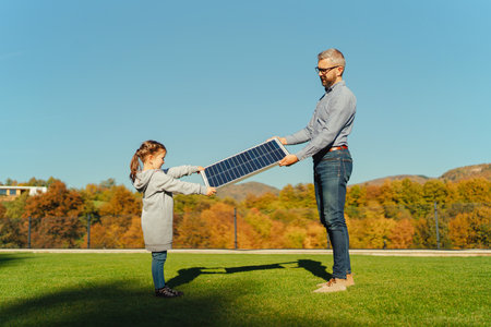 Father with his little daughter catching sun at solar panel,charging at their backyard. Alternative energy, saving resources and sustainable lifestyle concept.の写真素材