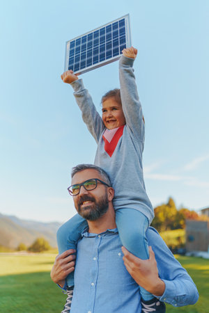 Father with his little daughter on piggyback, catching sun at solar panel,charging it at their...