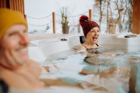 Senior couple in kintted cap enjoying together outdoor bathtub at their terrace during cold winter day.の写真素材
