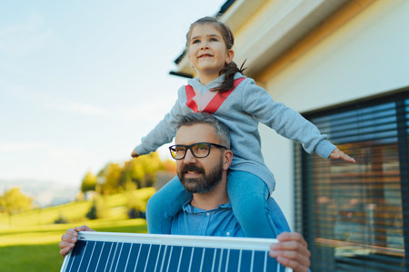 Father with his little daughter on piggyback, catching sun at solar panel,charging it at their backyard. Alternative energy, saving resources and sustainable lifestyle concept.の写真素材