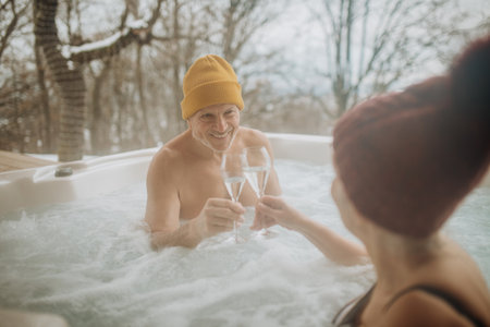 Senior couple in kintted cap enjoying together outdoor bathtub and clinking glasses at their terrace during cold winter day.の写真素材
