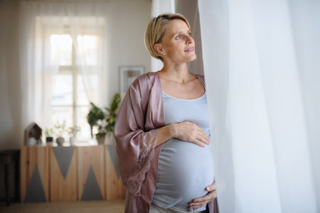Happy pregnant woman stroking her belly and looking out of the window.の写真素材