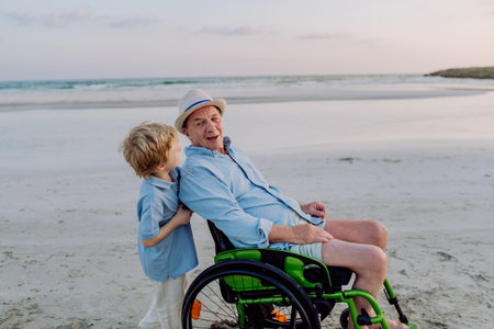 Little boy pushing his granfather on wheelchair, enjoying sea together.