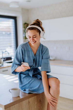 Young woman listening music, scrolling phone and enjoying cup of coffee at morning, in her kitchen.の写真素材