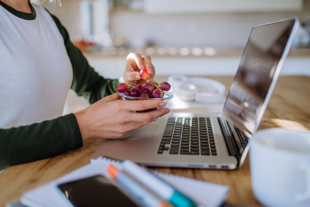 Side view of woman holding bowl with grapes above desk with computer, diary and smartphone....